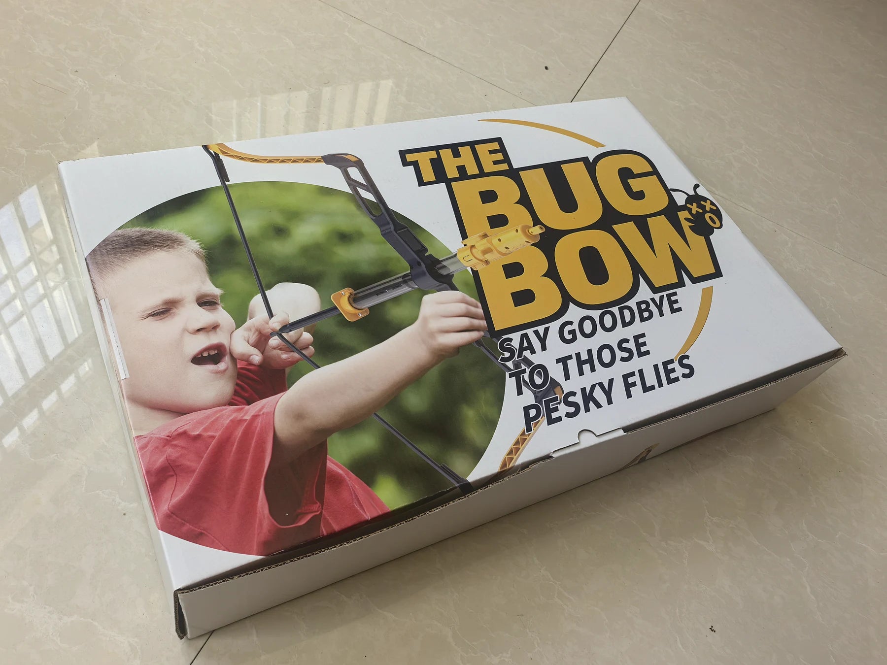 Physical BUG BOW retail box photographed on a table, showing a child aiming the Bug Bow with the logo and “Say goodbye to those pesky flies” tagline